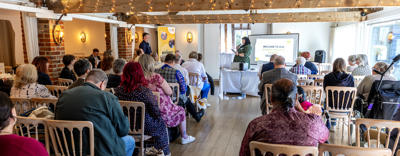 A photo from the back of the room, behind Audience members who are looking towards the stage during a presentation at the Resident Members Charter celebration event, April 2026.