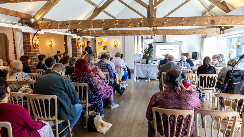 A photo from the back of the room, behind Audience members who are looking towards the stage during a presentation at the Resident Members Charter celebration event, April 2026.