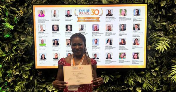 Mavis is stood in front of a screen detailing the 30 individuals featured in in Inside Housing Management 30. She is holding a certificate.