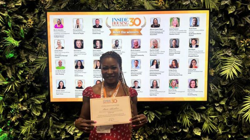 Mavis is stood in front of a screen detailing the 30 individuals featured in in Inside Housing Management 30. She is holding a certificate.