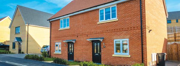 An Eastlight red-brick semi-detached home on a new housing development, with two front doors, white-framed windows and neatly maintained planting along the front. The property sits beside a paved driveway, with other homes visible in the background under a clear blue sky.