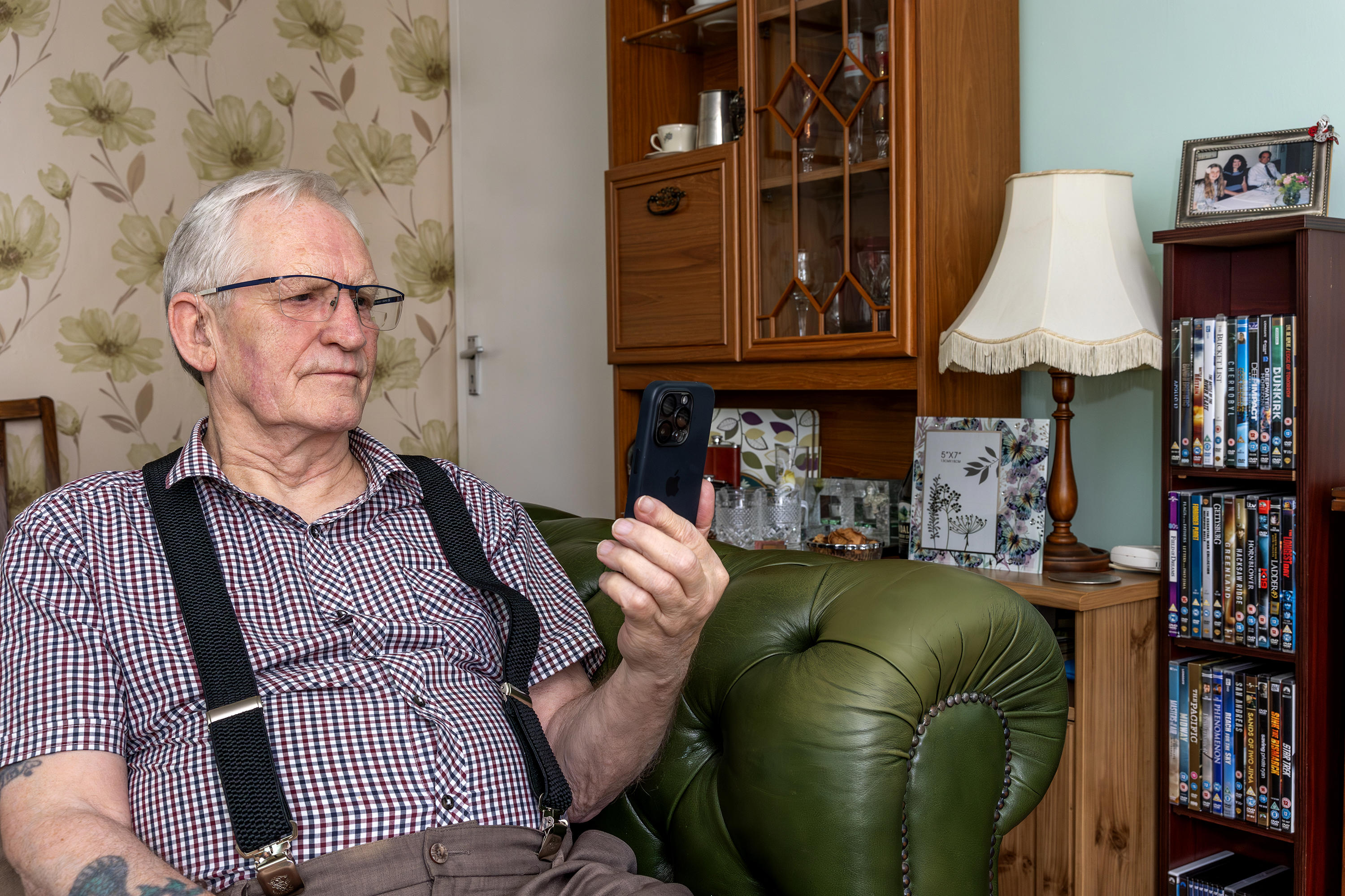An older man with grey hair and glasses is sat on his sofa in his home. He is holding up his mobile phone and looking at the screen.