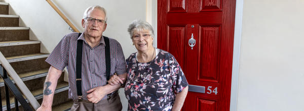 An older man and woman are stood together, with one of their arms linked, in front of their red front doors in a block of flats. The man is also leaning on the staircase to his right.