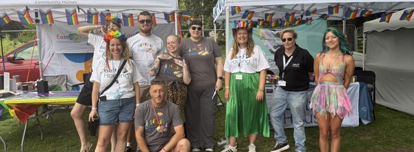 Eastlight Community Homes and Delta Housing staff stand together at an Essex Pride event beside rainbow-decorated information stalls, wearing EastPride T-shirts and colourful Pride accessories.