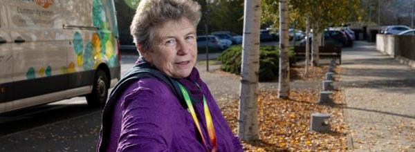 Eastlight's Chair of the Board, Hattie Llewelyn-Davies, sitting on a bench outside Eastlight House, Braintree, smiling at the camera. There are some trees and an Eastlight van in the background.  Hattie is wearing a purple jacket.