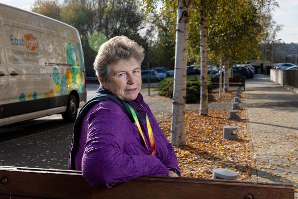 Eastlight's Chair of the Board, Hattie Llewelyn-Davies, sitting on a bench outside Eastlight House, Braintree, smiling at the camera. There are some trees and an Eastlight van in the background.  Hattie is wearing a purple jacket.