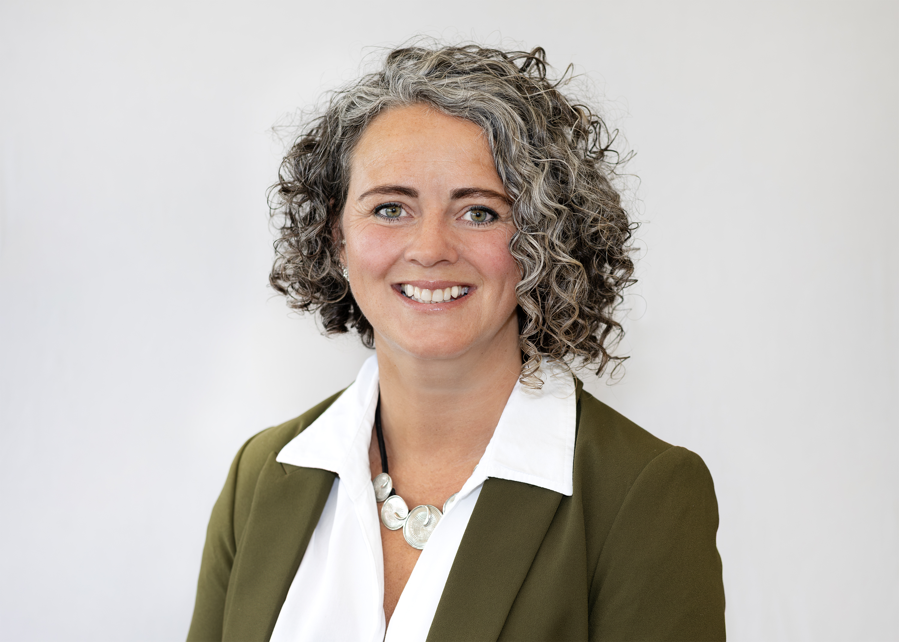 Jo Barrett is facing the camera and smiling. She has brown tight curly hair, greying at the front, and is wearing an open colour shirt and blazer.