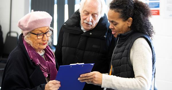 An older man and woman are stood next to each other and alongside a lady in a gilet. The older woman is wearing pink beret and is holding a blue clipboard. They are all looking at the clipboard.