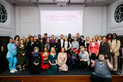 The full group of LCA Award Winners and Finalists up on stage together, smiling at the camera. The winners are sitting in the front row, holding their awards. The rest of the finalists are standing behind them.