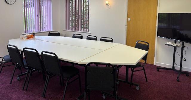 A large room with a large table and 10 chairs. There is a TV with stand in the corner.
