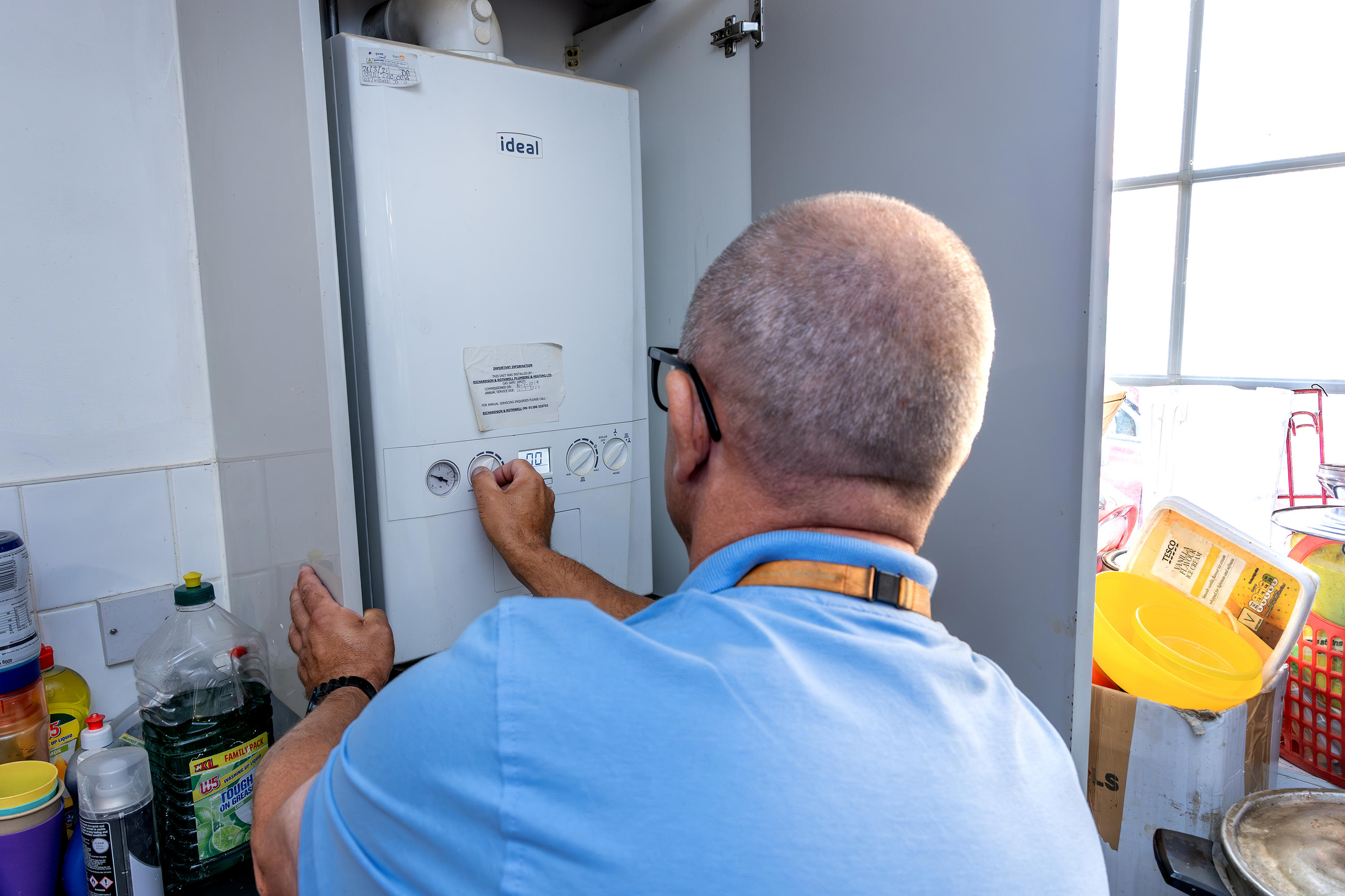  A member of Eastlight's Home Solutions Team carrying out a boiler check. He is photographed from behind and is wearing a blue top and an orange-coloured, Eastlight lanyard around his neck.