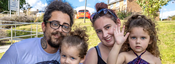 A man and woman are crouched down behind two young children. The family are all smiling at the camera.
