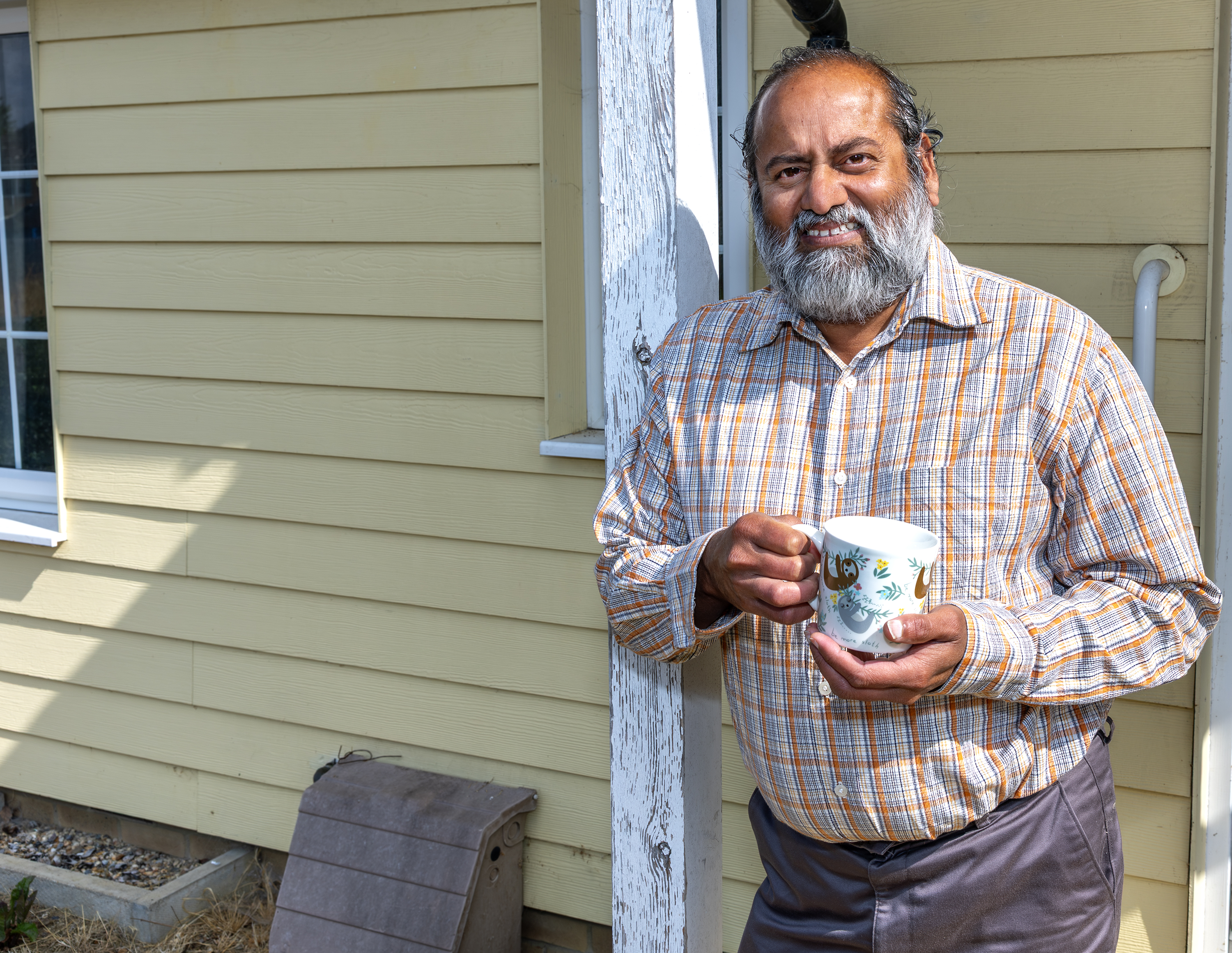 An Eastlight resident is leaning against a wooden post outside his home. He is holding a mug of tea.