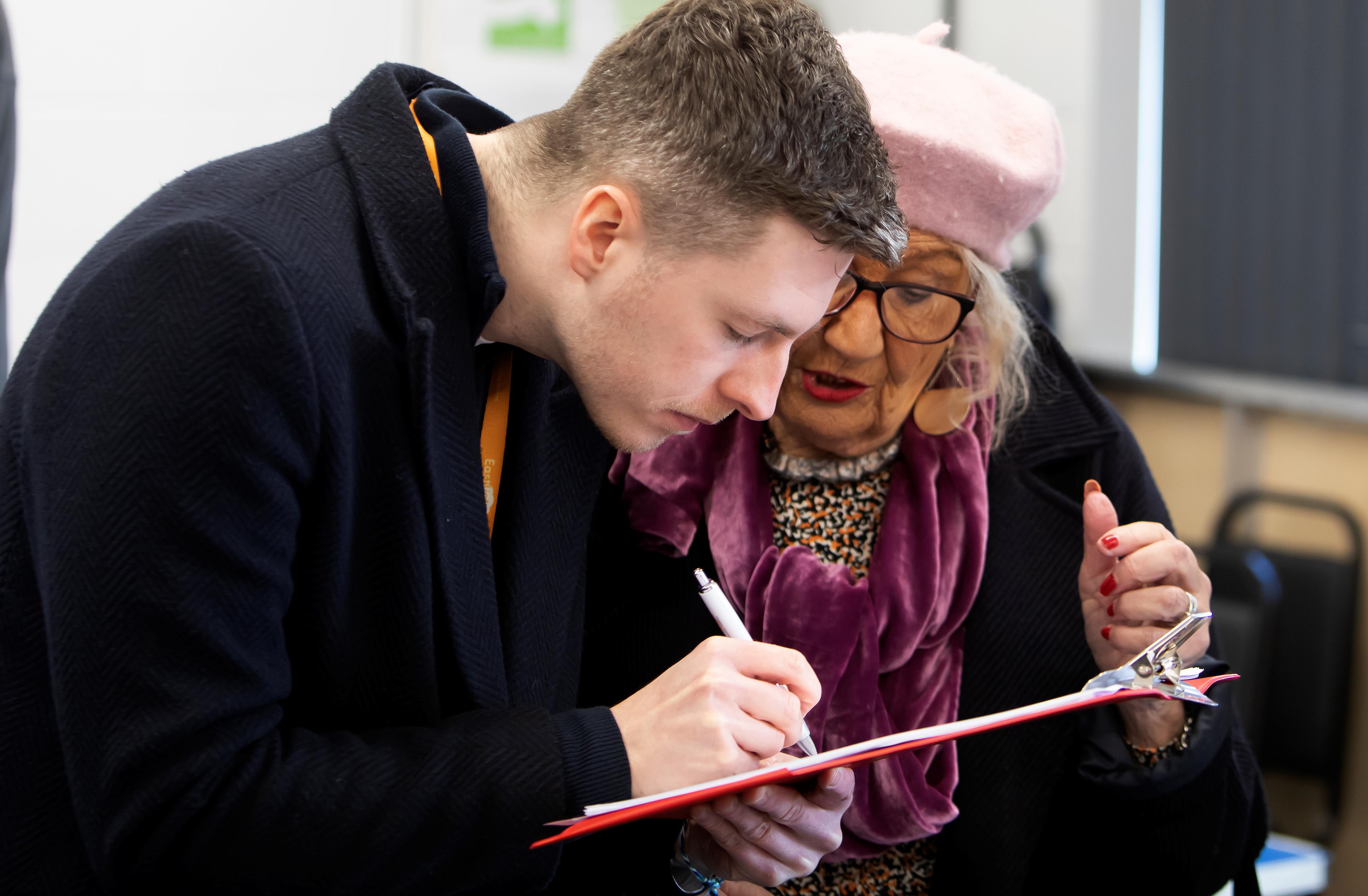 A man and a woman are stood together looking at a clipboard. The man is writing on the clipboard with a pen.