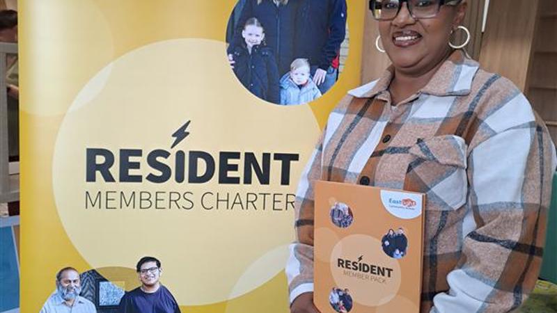 An Eastlight resident posing in front of a Resident Members Charter banner, holding her Resident Members Charter pack. She is smiling at the camera.
