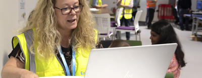 Eastlight Facilities Manager, Helen Symonds, is sat at a desk in front of a laptop. She is wearing a neon hi-vis vest and is holding a computer mouse in her right hand while looking at a laptop screen,