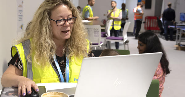 Eastlight Facilities Manager, Helen Symonds, is sat at a desk in front of a laptop. She is wearing a neon hi-vis vest and is holding a computer mouse in her right hand while looking at a laptop screen,