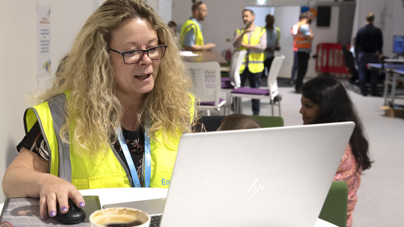Eastlight Facilities Manager, Helen Symonds, is sat at a desk in front of a laptop. She is wearing a neon hi-vis vest and is holding a computer mouse in her right hand while looking at a laptop screen,