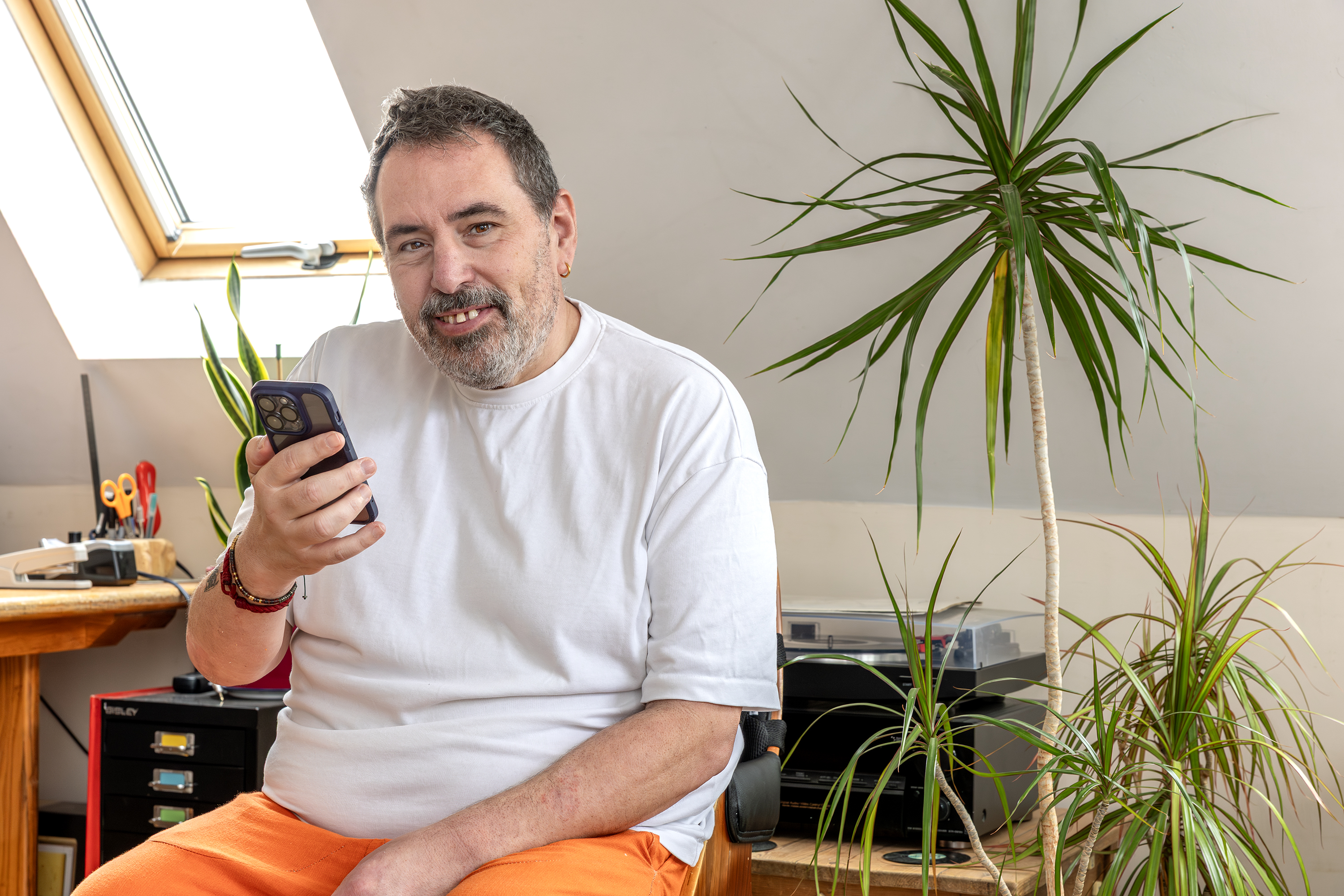 Eastlight resident and Customer Influence Committee member, Paul Hocker, sitting in his living room, holding a mobile phone, smiling at the camera. In the background are white walls, a number of houseplants and a stereo player on a small coffee table. 