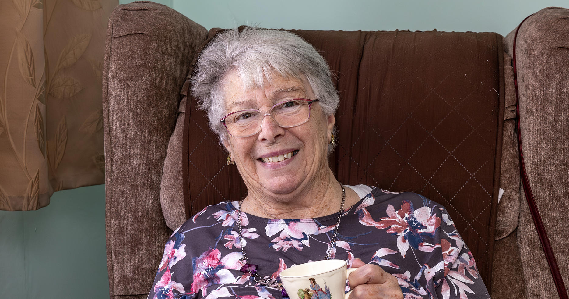 An older lady with glasses is holding a cup and saucer. She is smiling.