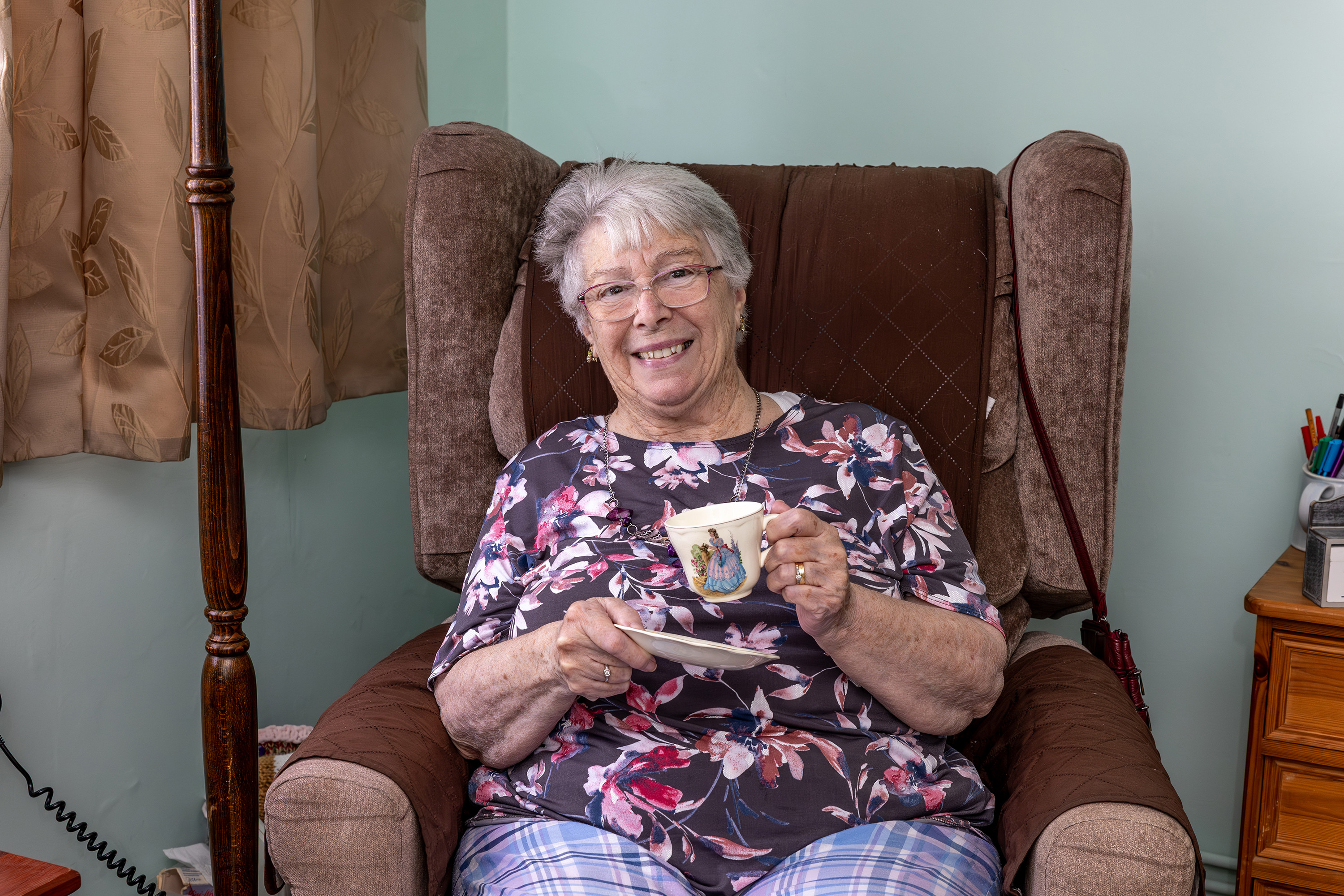 An older lady with glasses is holding a cup and saucer. She is smiling.