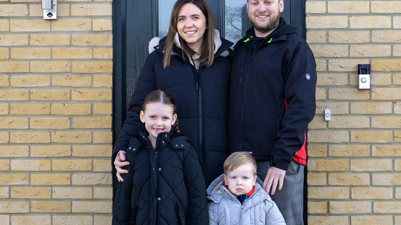 A family of Eastlight residents - including a Mum, Dad and two young children - standing on their front doorstep, smiling at the camera. The front door behind them is black, and the bricks are a light beige colour. There is a porch roof/cover over their heads. 