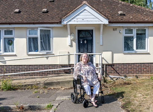 Eastlight resident, Tammy, is sat in her wheelchair outside her bungalow. There is a wheelchair ramp behind her than leads to the front door.