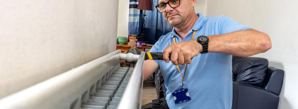 A member of Eastlight's Home Solutions Team working on a radiator inside a home, using a screwdriver. He has glasses and is wearing a blue top and an orange-coloured, Eastlight lanyard around his neck. In the background is a sofa and a doorway to another room in the house.