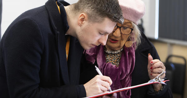 A man and a woman are stood together looking at a clipboard. The man is writing on the clipboard with a pen.