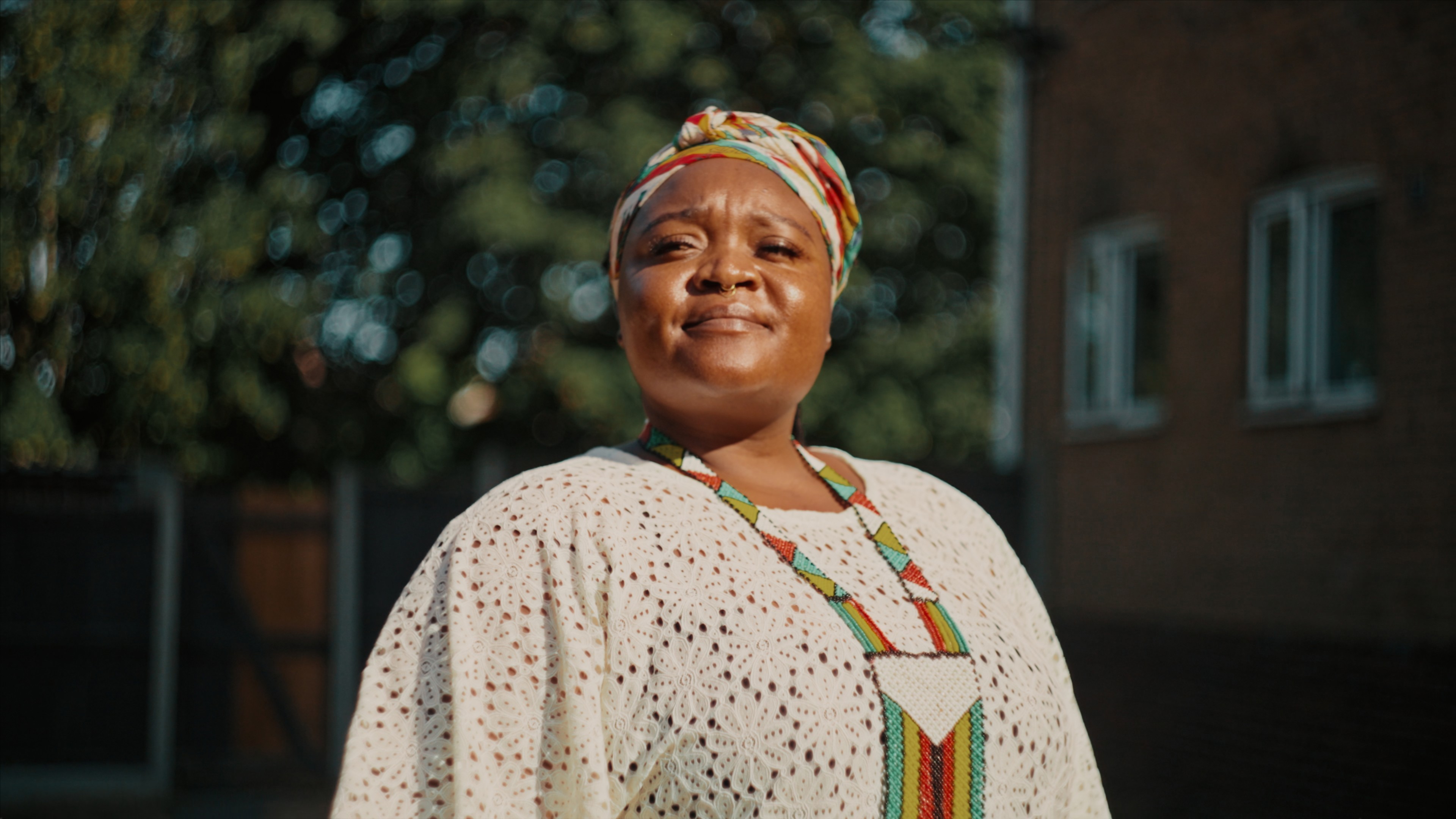 A woman wearing a white dress and Zimbabwean necklace and headscarf is standing proudly looking into the sun.
