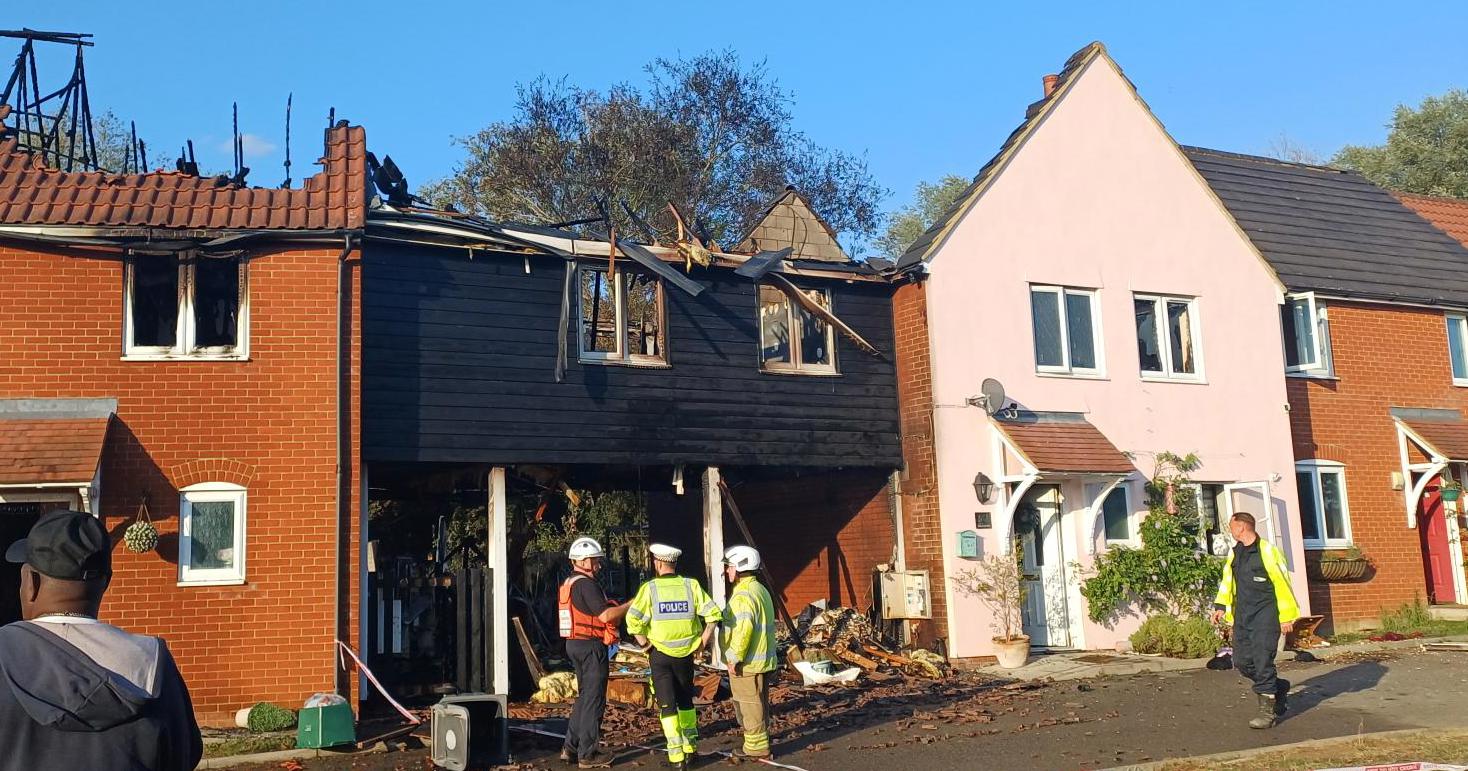 A row of terraced houses impacted by a fire. The roofs of the houses are missing. Police and firefighters are stood in front of the homes.