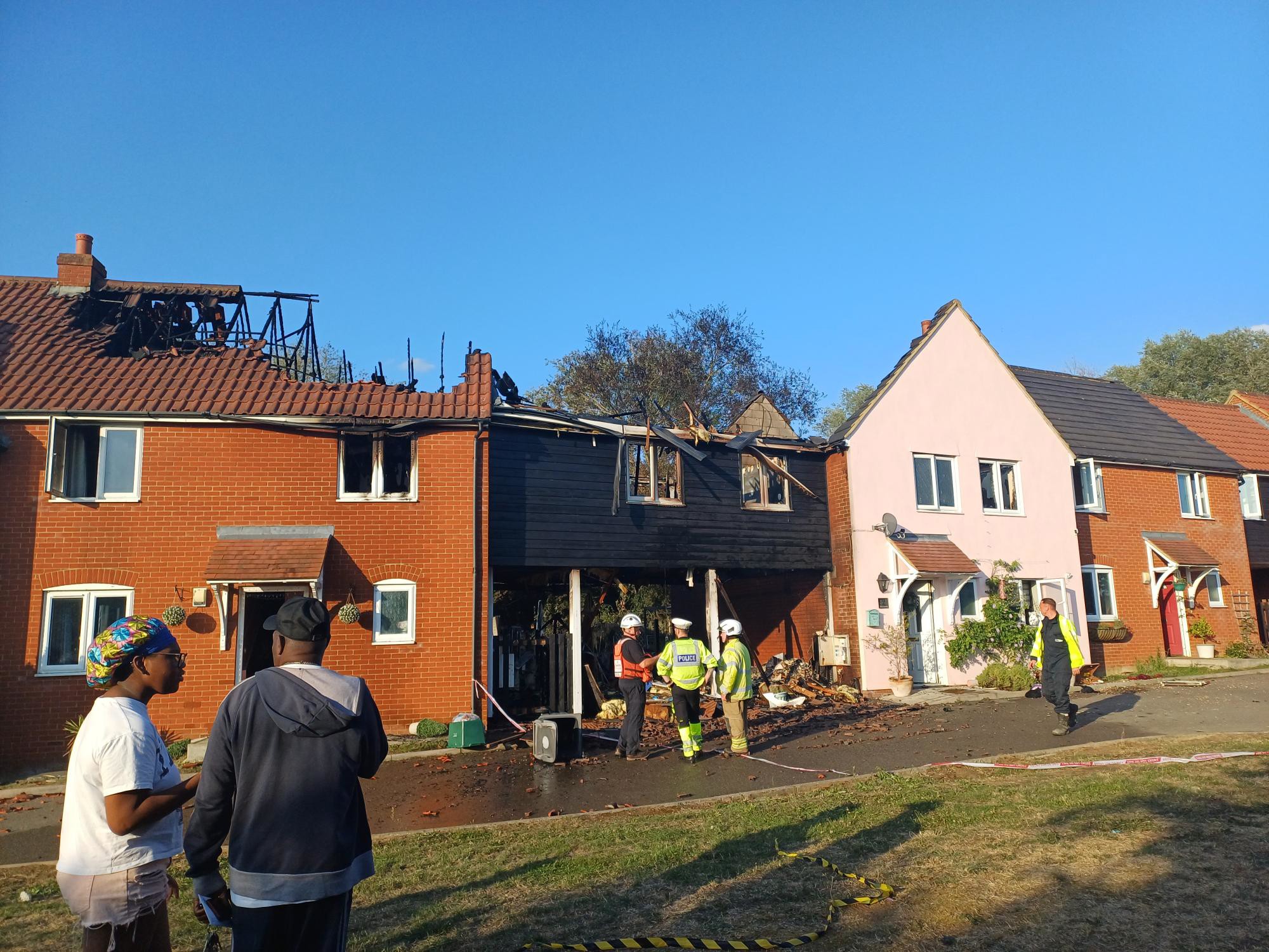 A row of terraced houses impacted by a fire. The roofs of the houses are missing. Police and firefighters are stood in front of the homes.