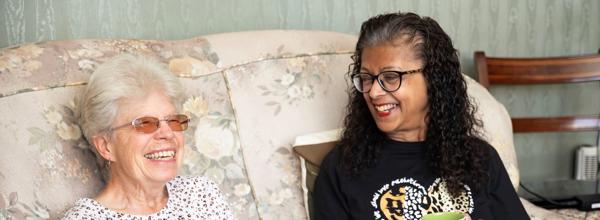 Two women sat on a sofa holding mugs of tea. They are both laughing and smiling.