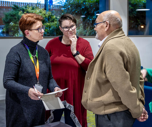 A man and two women are stood together in a group. They are all looking at each other, mid-conversation.