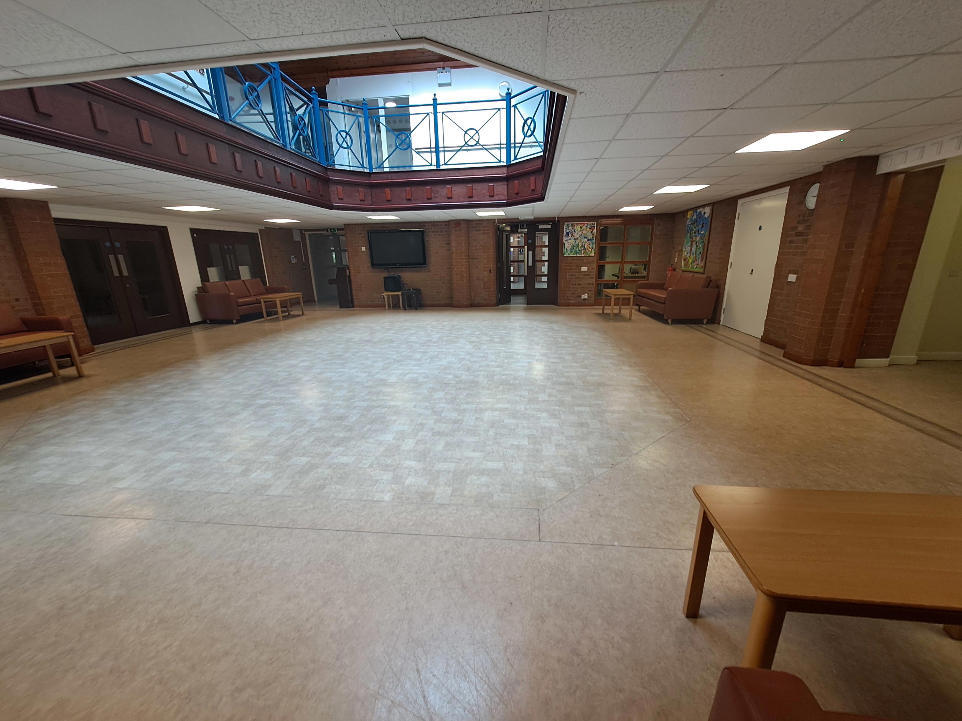 Large indoor atrium with an open central floor area, surrounded by exposed brick walls, brown sofas, and wooden side tables. A mezzanine balcony with blue metal railings overlooks the space from above through a polygon-shaped ceiling opening. The room is brightly lit by ceiling lights and natural light from upper windows, with doors, artwork, and a wall-mounted screen visible around the perimeter.
