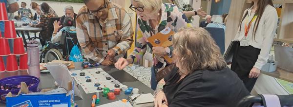 Eastliight residents gathering around a table at Eastlight House, Braintree, at the 2025 launch event for the Resident Members Charter. On the table are red cups in a pyramind shape, some large pieces of white paper and various coloured pens. 