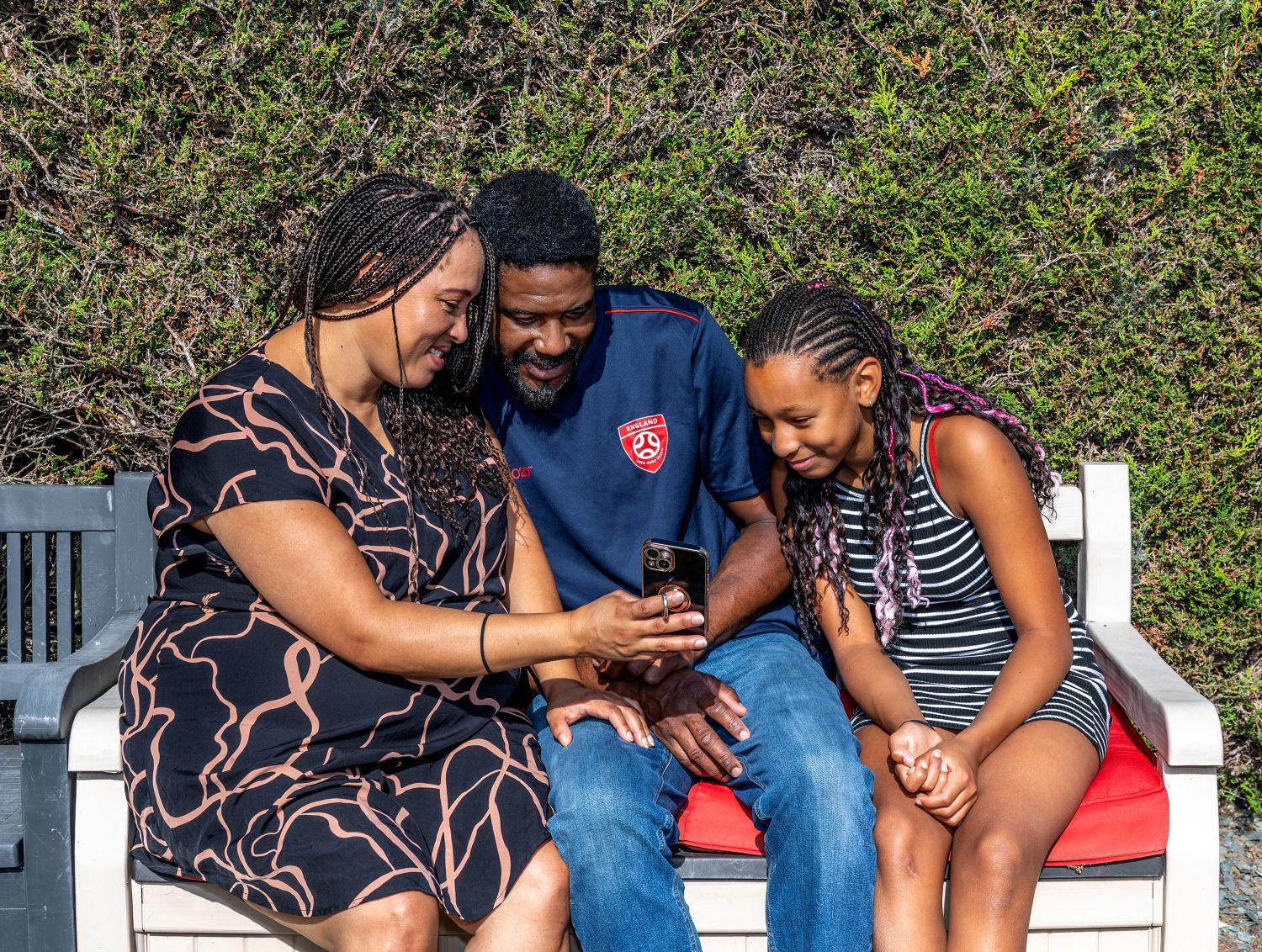 An Eastlight family, including (from left to right), a Mum, Dad and child all sitting outside on a bench, huddled over and smiling down at a mobile phone screen. There is some greenery (a bush) in the background. 