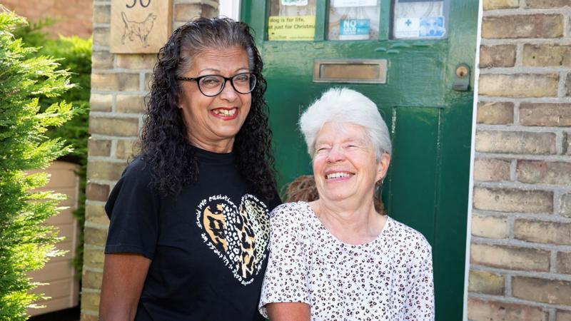 Eastlight residents Abaida and Joy, standing together on a front door step, smiling at the camera. The front door behind them is a dark green colour. To the left of the door is the house number 30. 