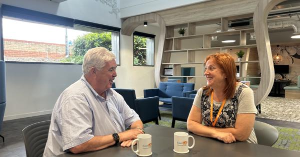 Former Eastlight Board member, Dale Butcher, sat with past Dale Butcher Work Experience Programme participant, Wendy Harris, in the downstairs cafe area of Eastlight House, Braintree. They are sitting at a table facing each other, smiling and chatting. There are two mugs of tea on the table, each with the Eastlight logo on them. The Eastlight offices are visible in the background.