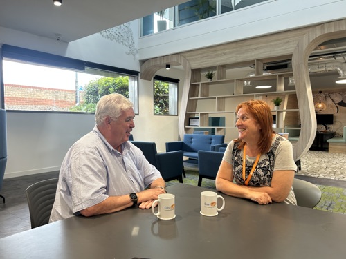 Former Eastlight Board member, Dale Butcher, sat with past Dale Butcher Work Experience Programme participant, Wendy Harris, in the downstairs cafe area of Eastlight House, Braintree. They are sitting at a table facing each other, smiling and chatting. There are two mugs of tea on the table, each with the Eastlight logo on them. The Eastlight offices are visible in the background.