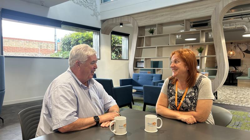 Former Eastlight Board member, Dale Butcher, sat with past Dale Butcher Work Experience Programme participant, Wendy Harris, in the downstairs cafe area of Eastlight House, Braintree. They are sitting at a table facing each other, smiling and chatting. There are two mugs of tea on the table, each with the Eastlight logo on them. The Eastlight offices are visible in the background.
