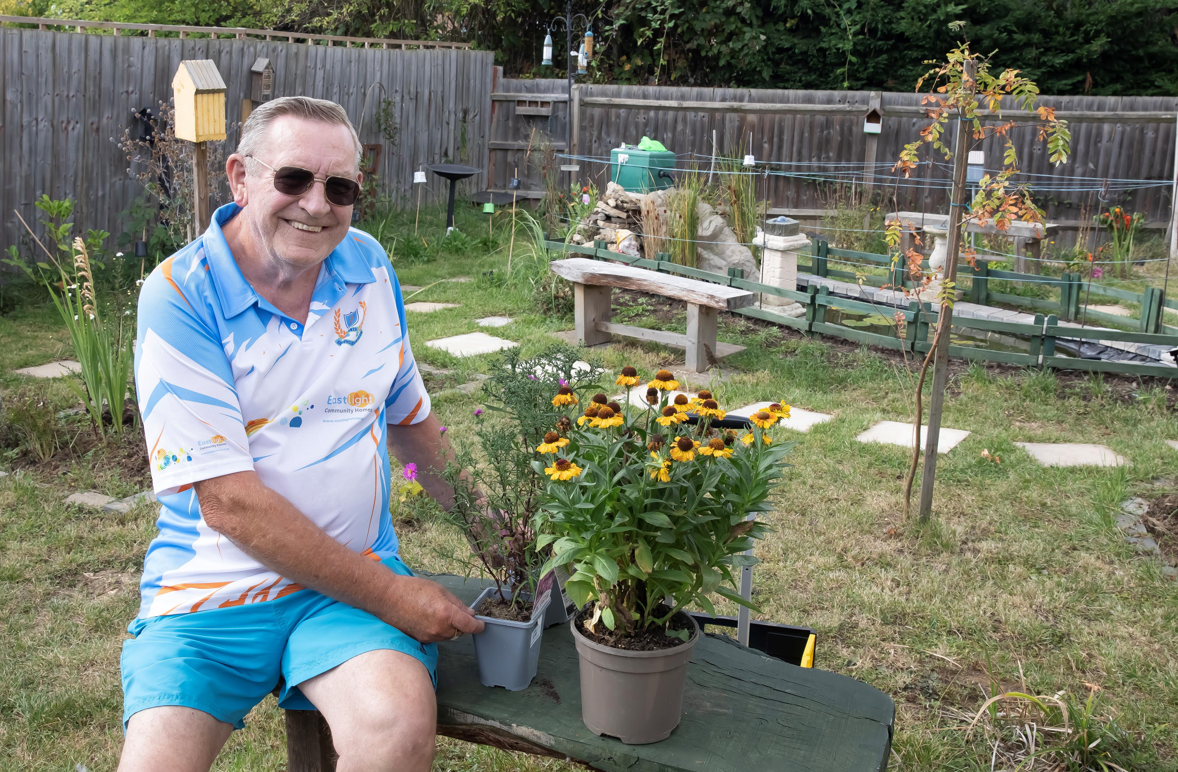 Jeff Spencer is sat on a bench in the community garden in Silver End. There are pots of flowers around him.