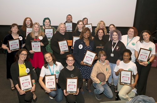 All four All In teams made up of 20 people stood together all each holding a certificate presented to them at the All In Ideas Festival in April 2023.